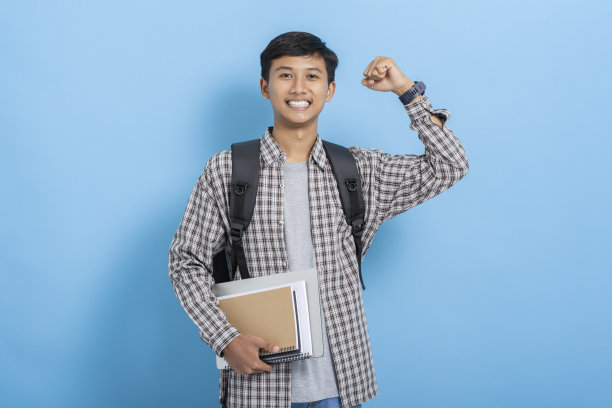 年轻的亚洲大学生背着书和背包，兴奋地庆祝成功，手臂在蓝色背景下孤立地举起图片下载