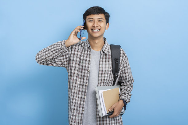 年轻的亚洲大学生背着书和背包打电话，在蓝色背景下孤立地微笑图片下载