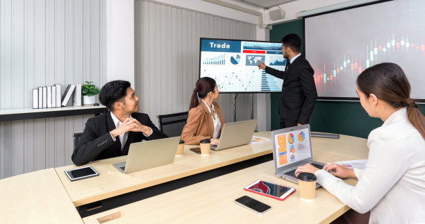 身穿黑色西装的商人指着大型数字显示器上的全球地图。企业高管团队在现代化的办公室开会，桌上有笔记本电脑、平板电脑、手机和咖啡。图片下载