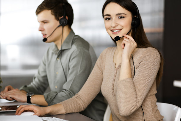 两名戴着耳机的年轻人坐在办公室的办公桌前与客户交谈。关注女性。呼叫中心接线员在工作图片下载