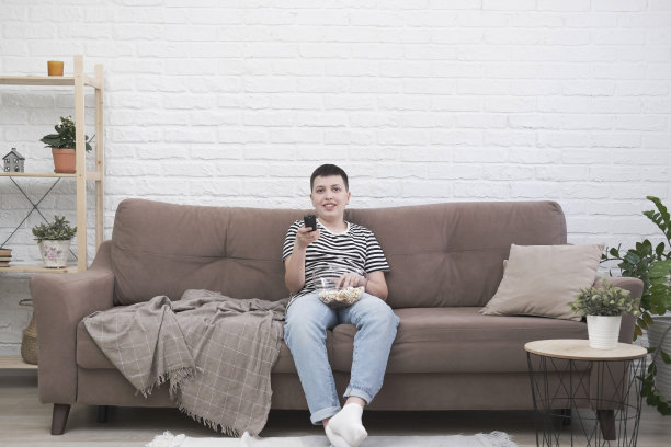 一个十几岁的男孩坐在沙发上，吃着爆米花，拿着控制面板，在明亮的客厅里看电视。图片下载