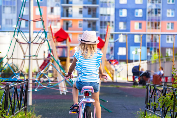 小女孩在粉红色的帽子骑自行车在户外操场的后视图。小金发孩子喜欢骑着小自行车在外面，夏天的时候。童年休闲活动概念。复制广告文字空间图片下载