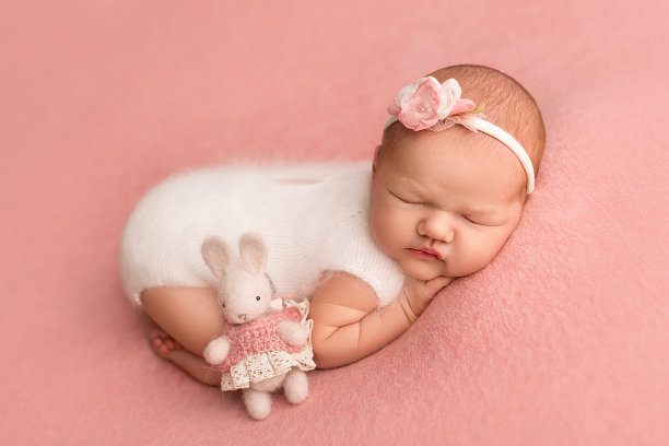 俯视图一个刚出生的女婴穿着白色工作服睡觉，头上缠着白色绷带。粉色背景上有一只针织的小白兔。新生婴儿7天，一周的美丽肖像。图片下载