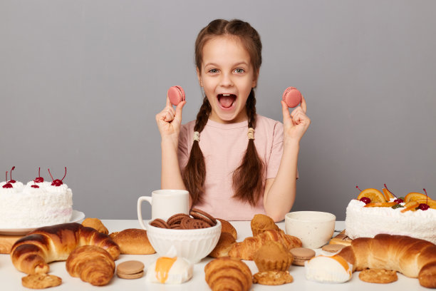 室内拍摄的惊喜非常快乐可爱的小女孩扎着辫子坐在桌子上满是自制糕点孤立在灰色背景，兴奋地尖叫，手里拿着马卡龙。图片下载