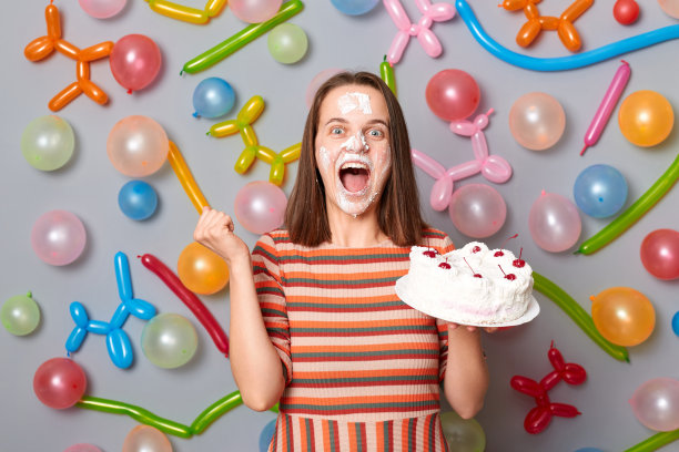 生日快乐的女人接受最好的朋友的祝贺把蛋糕放在脸上涂满奶油快乐地尖叫，张着嘴，拿着气球靠墙站着。图片下载