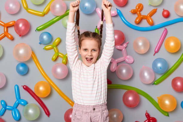 疯狂的生日小女孩，扎着辫子，穿着休闲的衣服，站在灰色的墙上，有彩色的气球，有一个有趣的脸，把她的辫子拉起来。图片下载