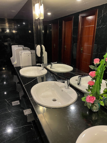 酒店公共浴室的白色瓷水槽，金属水龙头和插头孔，黑色大理石墙壁和地板，红木门，架子上的人造玫瑰，背景是小便池图片下载