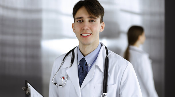 友好的年轻男性医生站在现代诊所夹板。女同事在医生的后台忙着填写用药记录表。医学的概念图片下载