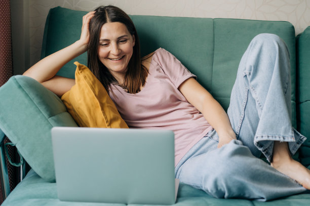 四十多岁的快乐微笑的女人坐在家里的沙发上用笔记本电脑工作。图片下载