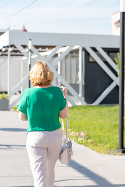 后视图的时尚苗条的女人在绿色衬衫，裤子和手提包走在街道上图片下载