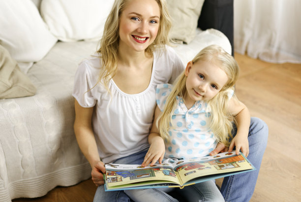 幸福的家庭。年轻的金发妈妈坐在木地板上给她可爱的女儿读书。母亲的概念图片下载