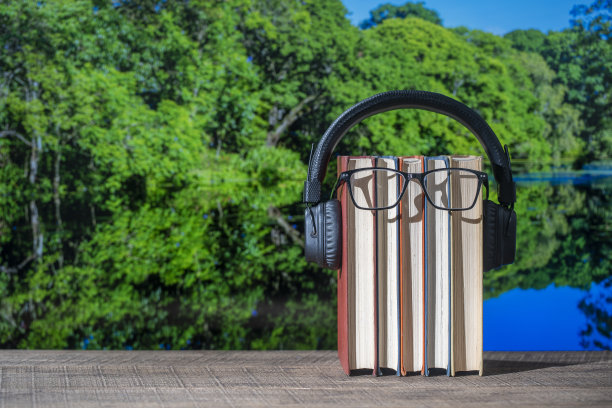 一套书，耳机和眼镜在木桌与湖水和森林背景在一个阳光明媚的夏日，特写。有声读物和教育理念图片下载