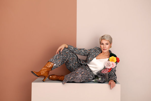 时尚成熟的女人躺在工作室的棕色和米色背景。中年时尚模特，短发，穿着时髦的裤装，带跟的皮革短靴图片下载