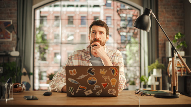 专业的创意男子坐在办公桌在家庭办公室工作室，在笔记本电脑上工作。专家思考一封好的邮件回复。在家工作的男性员工寻找灵感图片下载