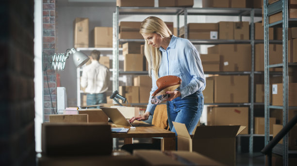 库存经理包装一个时尚的自行车座位卖给客户在线。准备一个小纸板包裹邮资。金发女小企业主在仓库工作。图片下载