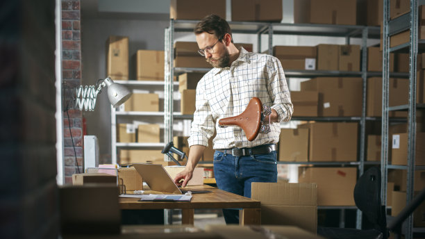 小企业主包装复古自行车座位卖给客户在线。准备一个小纸板箱邮资。在仓库工作的男性库存经理。图片下载