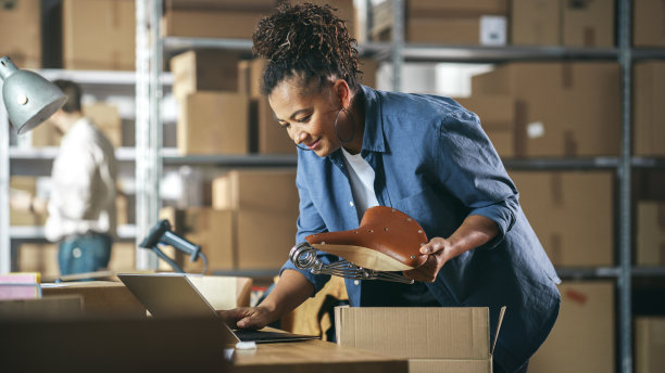 小企业主包装一个时尚的自行车座位卖给客户在线。准备一个小纸板箱邮资。在仓库工作的黑人女性库存经理。图片下载