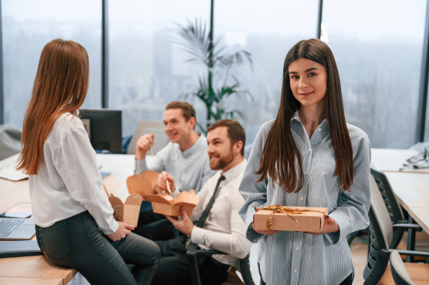 女人站在那里，手里拿着节日礼物。一群同事在办公室吃环保盒里的食物图片下载