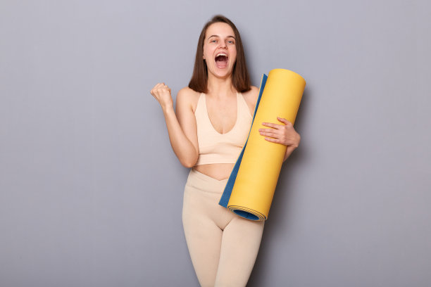 照片中非常快乐的运动女性穿着运动服，手拿瑜伽垫，在灰色背景上隔离，握紧拳头，庆祝训练后的优异成绩。图片下载