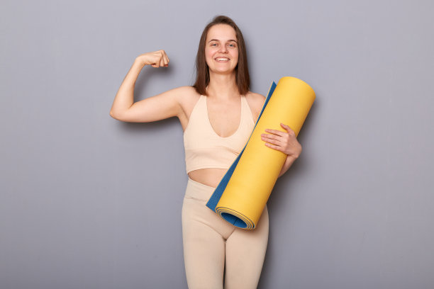 肖像满意自豪的运动员在米色运动服抱着健身垫，展示她的肱二头肌，在运动训练后展示她的力量。图片下载