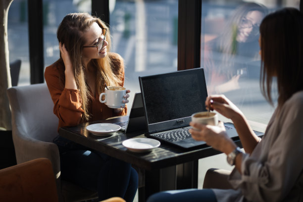 两个年轻的女商人在咖啡馆里一对一地开会。朋友们下班后聊八卦，坐在靠窗的桌子边喝咖啡。图片下载