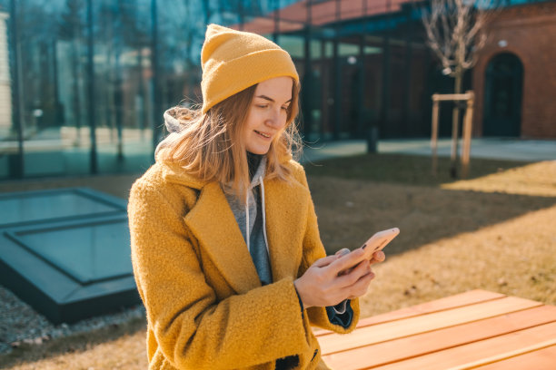 照片金发快乐的学生女子20多岁手持电话穿着黄色外套和帽子坐在城市春天公园户外休息使用手机在线聊天的长椅上。图片下载