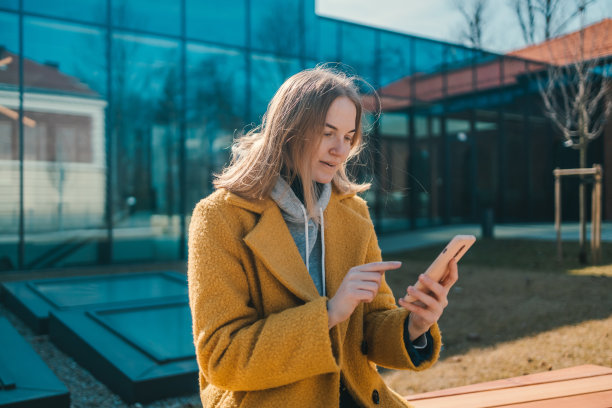 照片金发快乐的学生女子20岁手持手机穿着黄色外套，坐在城市春天公园户外休息使用手机在线聊天的长椅上。图片下载