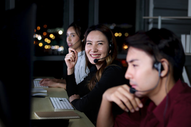 快乐微笑的亚洲女性在呼叫中心工作，与队友同事一起通宵工作，夜班客服人员戴着麦克风耳机使用电脑为客户提供支持图片下载