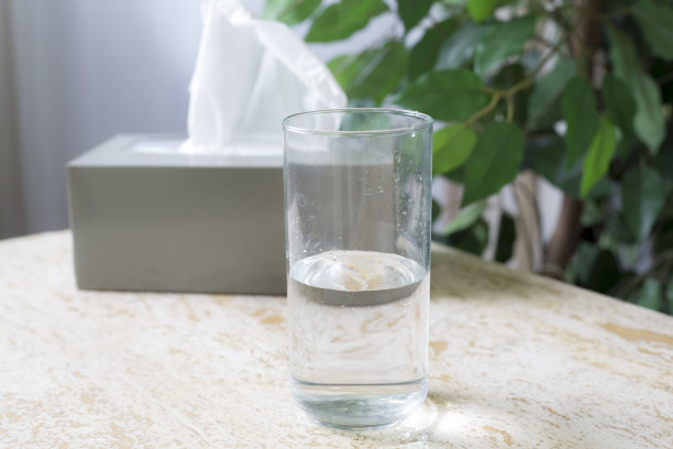 一盒纸巾放在桌子上，背景是绿色植物。纸巾盒旁边放着一杯水。图片下载