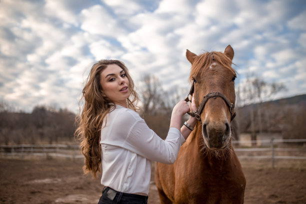 快乐的年轻女子站在她的棕色马旁边看着它。她握着缰绳。背景是白色的栅栏和美丽的天空和白云。图片下载
