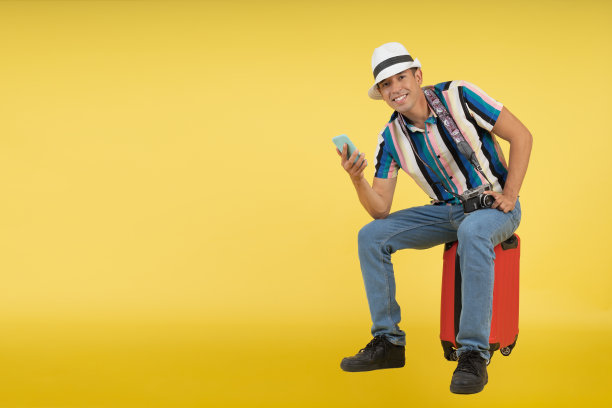 年轻的旅行者，坐在行李箱上，拿着手机，微笑着看着镜头。图片下载