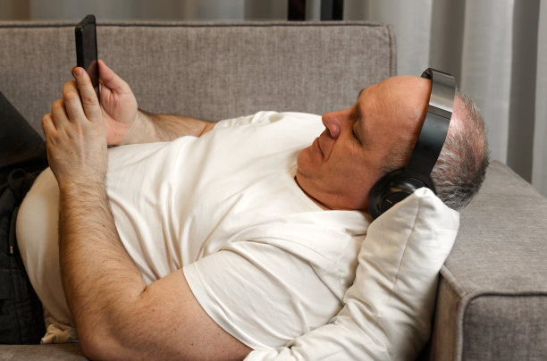 一个中年男人躺在沙发上，戴着耳机听音乐，看手机图片下载