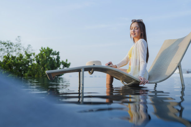 微笑开朗的亚洲成年女子穿着比基尼放松休闲日光浴在泳池长凳屋顶游泳池城市城市生活方式，暑假旅游，女子坐在长椅上幸福和平无忧无虑图片下载