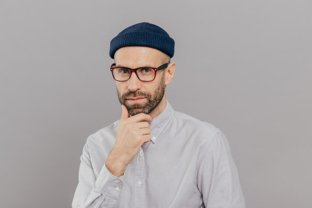 蓝色眼睛的成年男性，托着下巴，专注地看着镜头，思考着出路或解决方案，戴着眼镜，帽子和正装衬衫，在灰色的工作室墙壁上模特。商人在室内拍照图片下载
