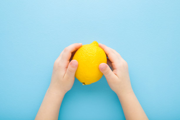 淡蓝色的桌子背景上，小孩子的手拿着黄色的柠檬。柔和的颜色。新鲜水果。特写镜头。视角镜头。自顶向下视图。图片下载