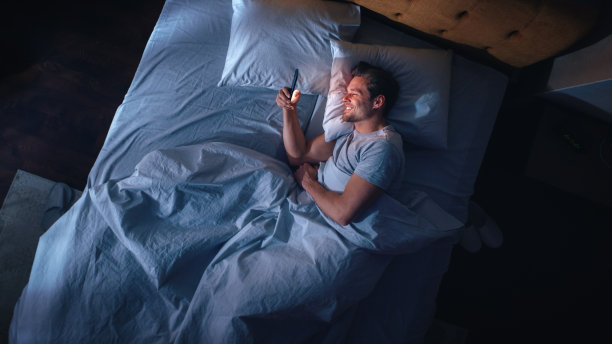 俯视图公寓卧室:英俊的年轻男子晚上在床上使用智能手机。微笑的家伙浏览社交媒体，约会应用程序，远程工作软件，在手机上做网上购物图片下载