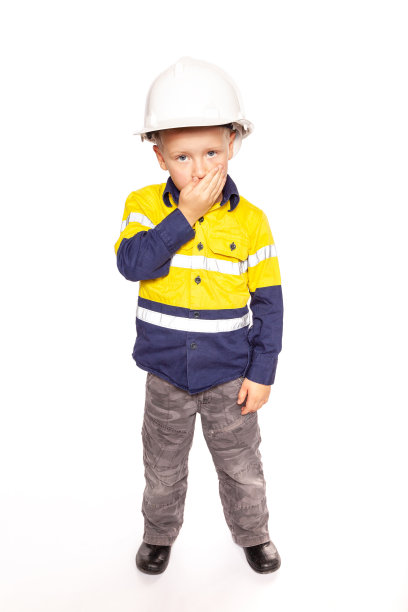 年轻的金发白人男孩手捂嘴，一副震惊的样子，角色扮演一名建筑工人主管，穿着黄蓝相间的hi-viz衬衫，靴子，戴着白色安全帽。图片下载