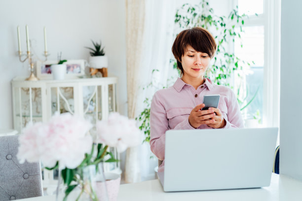 女人用笔记本电脑在家里休息时使用手机。舒适的工作空间，新鲜的牡丹花和一杯热饮放在白色的桌子上，室内背景明亮。远程工作。图片下载