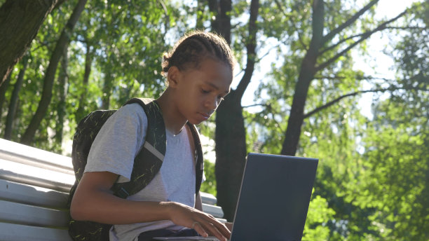 一个非裔美国少年拿着笔记本电脑坐在公园的长椅上。图片下载
