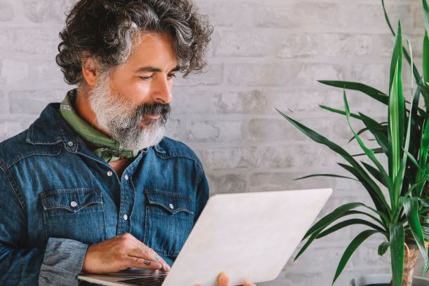 大胡子成年男子，坐在椅子上使用笔记本电脑，背景是一堵墙和一棵植物图片下载