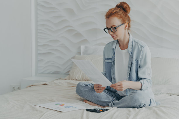 年轻女子盘腿坐在舒适的床上检查财务文件计算家庭预算图片下载
