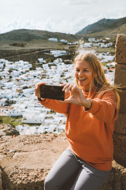 旅行博主女子用智能手机拍摄自拍照片，网红女孩在希腊观光林多斯镇鸟瞰罗德岛夏季户外旅行图片下载