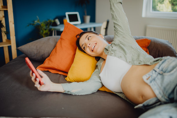 微笑的孕妇躺在床上使用智能手机图片下载