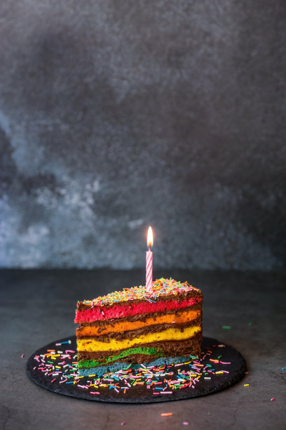 彩虹生日蛋糕装饰着喜庆的蜡烛和五彩纸屑。糖果。五彩缤纷的甜点，在黑色的背景上有彩虹的颜色。图片下载
