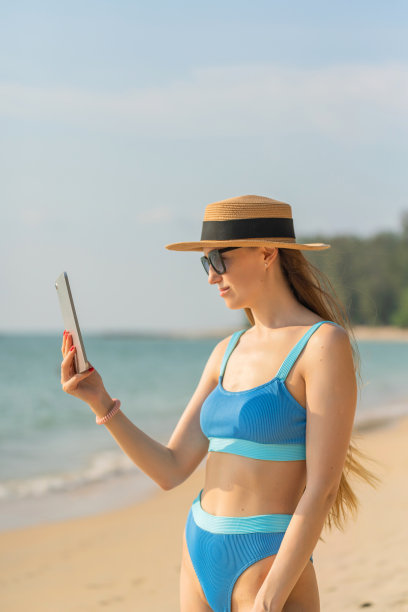 数字生活。穿着比基尼的美丽白人女子使用她的数字平板电脑与她的团队一起工作，并在海滩上与她的朋友愉快地交流图片下载