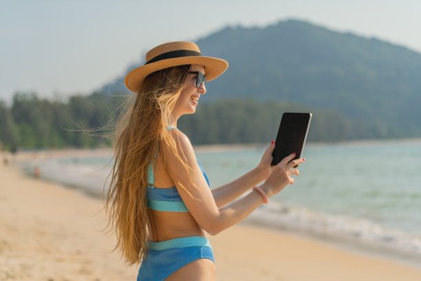 数字生活。穿着比基尼的美丽白人女子使用她的数字平板电脑与她的团队一起工作，并在海滩上与她的朋友愉快地交流图片下载