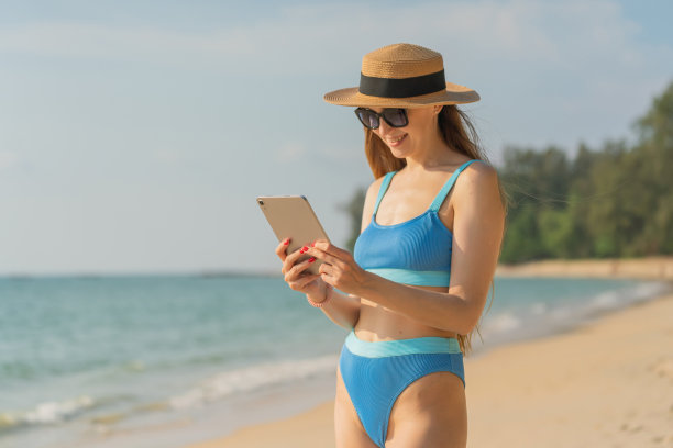 数字生活。穿着比基尼的美丽白人女子使用她的数字平板电脑与她的团队一起工作，并在海滩上与她的朋友愉快地交流图片下载
