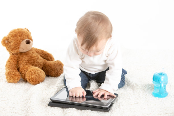 白色背景的工作室照片，一个婴儿触摸平板电脑的屏幕，旁边有一只泰迪熊图片下载