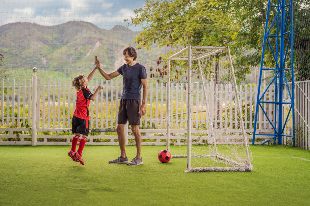穿着红色足球服的小男孩和他的教练或父亲在户外踢足球。活泼的孩子与孩子或父亲一起运动，微笑的快乐男孩在夏天玩得开心图片下载
