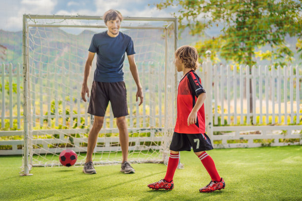 穿着红色足球服的小男孩和他的教练或父亲在户外踢足球。活泼的孩子与孩子或父亲一起运动，微笑的快乐男孩在夏天玩得开心图片下载